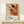 May include: Abstract expressionist artwork poster in light wood frame leaning against white wall on pale wooden surface. Features dynamic paint splatters, drips and brushstrokes in orange, red, black, yellow and green on cream background. Exhibition text below artwork reads "Jackson Pollock January 10-20, 1956 8th Avenue, New York City". Small white wrapped gift with twine and evergreen sprig sits in front of frame.