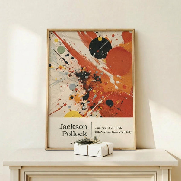 May include: Abstract expressionist artwork poster in light wood frame leaning against white wall on pale wooden surface. Features dynamic paint splatters, drips and brushstrokes in orange, red, black, yellow and green on cream background. Exhibition text below artwork reads "Jackson Pollock January 10-20, 1956 8th Avenue, New York City". Small white wrapped gift with twine and evergreen sprig sits in front of frame.