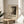 May include: Framed poster with abstract black and white splattered artwork displayed on round wooden side table next to beige upholstered chair. Poster features "POLLOCK" text and exhibition details dated 2030. Natural lighting creates soft shadows on white wall. Green plant leaves visible in corner. Minimalist modern interior styling with neutral tones throughout the scene.