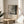 May include: Framed poster with abstract black and gray splattered artwork in the upper portion and white text area below, displayed on a round wooden side table next to a beige upholstered chair. Natural sunlight streams across the neutral-toned room, with a houseplant visible in the corner. The poster features a thin wooden frame and museum-style text layout.