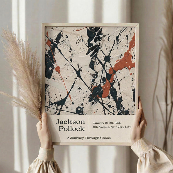 May include: Person holding framed abstract art print featuring black, orange, and beige paint splatters and drips on cream background. Wooden frame with exhibition-style text below artwork. Dried pampas grass visible to left. Soft natural lighting against neutral curtained background.