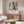 a modern living room with a large abstract painting hanging on the wall above a wooden cabinet. The painting has a mix of black, white, and orange colors, creating a striking visual contrast. The room also includes a white sofa, a round glass coffee table, and a lamp on a side table.