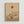May include: Minimalist art print in light wood frame on white wall featuring beige background with red-orange sun, geometric circles, bare branching tree with sparse foliage, and vertical line creating symmetrical composition in warm earth tones