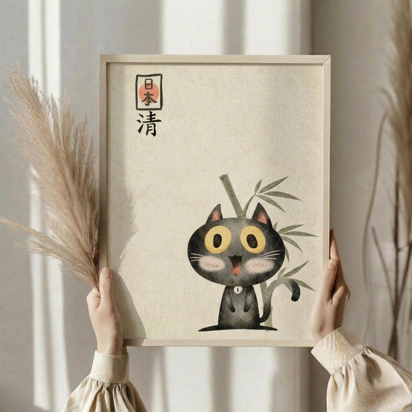 May include: Person holding framed artwork showing cute cartoon black cat with large yellow eyes and pink cheeks sitting next to green bamboo stalks, with Japanese characters above. Light wooden frame against cream textured background. Other hand holds dried pampas grass stems. Soft neutral lighting and beige clothing visible.