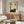 May include: Dark tabby cat resting in front of framed Asian-inspired artwork featuring green bamboo stalks and Japanese characters on cream background. Modern living room setting with walnut sideboard, white mushroom table lamp, cream curved chair, glass coffee table, and wooden floor lamp. Minimalist decor with natural wood tones and soft lighting.