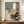 May include: Framed abstract artwork featuring geometric shapes in sage green, cream, and terracotta tones with black pine tree silhouette overlaid on composition, displayed on wooden credenza beside white ceramic vase and stacked books in neutral-toned room with white paneled walls