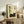May include: Framed Japanese-style artwork featuring a dark gray cat resting beside green bamboo stalks on cream background with black characters in upper right corner, displayed on rustic wooden console table alongside fern plant, books, ceramic vase, and brown leather chair in modern living room setting with white walls and natural lighting.