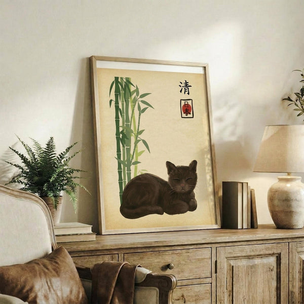 May include: Framed Japanese-style artwork featuring a dark gray cat resting beside green bamboo stalks on cream background with black characters in upper right corner, displayed on rustic wooden console table alongside fern plant, books, ceramic vase, and brown leather chair in modern living room setting with white walls and natural lighting.