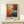 May include: Abstract expressionist artwork poster in wooden frame leaning against white wall on light wood surface. Vibrant painting features explosive splashes and drips of orange, red, blue, black and white paint with dynamic movement radiating from center. Text reads "POLLOCK Expressionist Art Movement" below colorful composition. Small white wrapped gift with green sprig sits in front of frame.