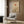 May include: Minimalist Japanese-inspired poster featuring a silhouetted tree beneath a red sun and beige moon on cream background, displayed in white frame on wood-paneled wall above beige upholstered armchair with geometric patterned pillow, alongside brass floor lamp and dark round side table with open book