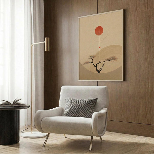 May include: Minimalist Japanese-inspired poster featuring a silhouetted tree beneath a red sun and beige moon on cream background, displayed in white frame on wood-paneled wall above beige upholstered armchair with geometric patterned pillow, alongside brass floor lamp and dark round side table with open book