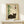 May include: Framed Japanese-style artwork featuring a sleeping dark gray cat beside green bamboo stalks, with black Japanese characters and red stamp seal on cream background. Displayed on light wood shelf with small white wrapped gift. Natural lighting creates soft shadows on white wall.