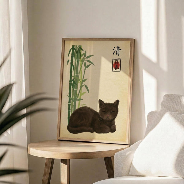 May include: Framed poster displayed on wooden round side table next to beige upholstered chair in sunlit room. Artwork features dark tabby cat resting beside green bamboo stalks on cream background with Japanese characters and small red rectangular element. Natural wood frame complements modern minimalist decor with plant shadows visible on white wall.