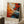 May include: Abstract expressionist artwork poster with vibrant orange, red, blue and white paint splatters and drips in wooden frame, leaning against white wall next to coffee cup on wooden block and cream textured blanket on white fluffy rug