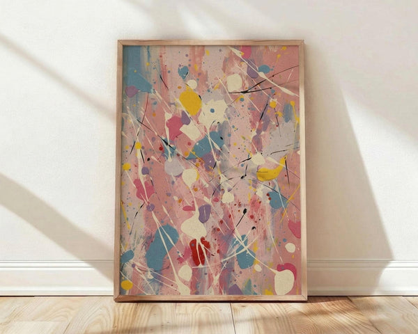 May include: Abstract expressionist style artwork in wooden frame leaning against white wall on light wood floor. Colorful paint splatters and drips in pink, blue, yellow, red, and cream create dynamic composition with black linear elements and organic shapes scattered across canvas. Soft natural lighting casts shadows on textured wall surface.