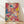 May include: Abstract expressionist artwork with vibrant paint splatters and drips in blue, pink, yellow, and red colors displayed in a light wooden frame, leaning against a white wall on light wood flooring with natural sunlight casting diagonal shadows across the scene.