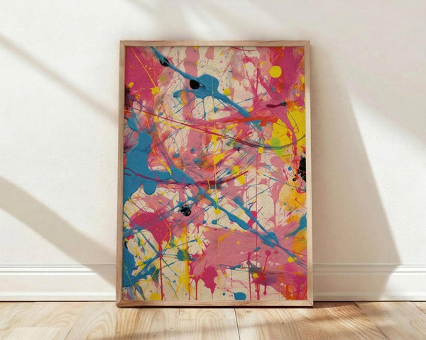May include: Abstract expressionist artwork with vibrant paint splatters and drips in blue, pink, yellow, and red colors displayed in a light wooden frame, leaning against a white wall on light wood flooring with natural sunlight casting diagonal shadows across the scene.