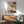May include: Framed Japanese-inspired artwork featuring a lone tree silhouette with sparse branches against a beige background, red circle representing sun in upper right, geometric black lines creating grid pattern overlay. Displayed above wooden sideboard with white ceramic lamp, next to sage green mid-century armchair with wooden arms. Warm sunlight creates shadows across white wall and wooden furniture in modern living room setting.