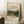 May include: Framed vintage-style museum poster featuring impressionist landscape painting of rolling green hills, country village, and cloudy sky. The artwork shows rural French countryside with stone path and scattered buildings. Poster has cream background with black text reading "The MET museum" and "CAMILLE Pissarro". Frame leans against white wall corner next to wooden side table with white coffee cup and saucer, positioned on textured cream rug beside beige fabric.