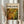 May include: Person in cream ruffled blouse holding framed abstract art print with swirling green, yellow, and orange paint splatters and circular patterns. Light wood frame against beige curtain backdrop with dried wheat stems visible on left side.