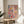 May include: Abstract expressionist artwork with vibrant paint splatters and drips in pink, blue, yellow, and red colors, displayed in thin wooden frame on round natural wood side table next to beige upholstered chair. Sunlight streams across white wall creating geometric shadows, with green plant leaves visible in corner of modern minimalist room setting.