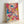 May include: Abstract expressionist style artwork in light wood frame leaning against white wall. Vibrant paint splatters and drips in bright blue, hot pink, yellow, and white create dynamic layered composition. Energetic brushstrokes and paint drips overlap across cream background, showing textured paint application typical of action painting technique.