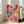 May include: Large framed abstract art print with vibrant paint splatters and drips in hot pink, turquoise blue, bright yellow, and orange colors, displayed in white frame leaning against wall next to beige upholstered chair in bright living room with white walls and large windows
