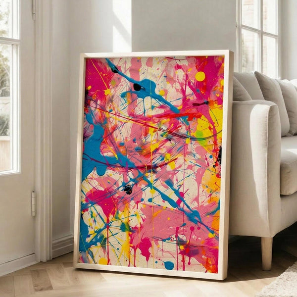 May include: Large framed abstract art print with vibrant paint splatters and drips in hot pink, turquoise blue, bright yellow, and orange colors, displayed in white frame leaning against wall next to beige upholstered chair in bright living room with white walls and large windows