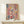 May include: Abstract expressionist style artwork with pink background featuring colorful paint splatters and drips in yellow, blue, red, and white with black lines throughout, displayed in thin wooden frame on light wood console table next to small wrapped white gift box with evergreen sprig, against cream wall