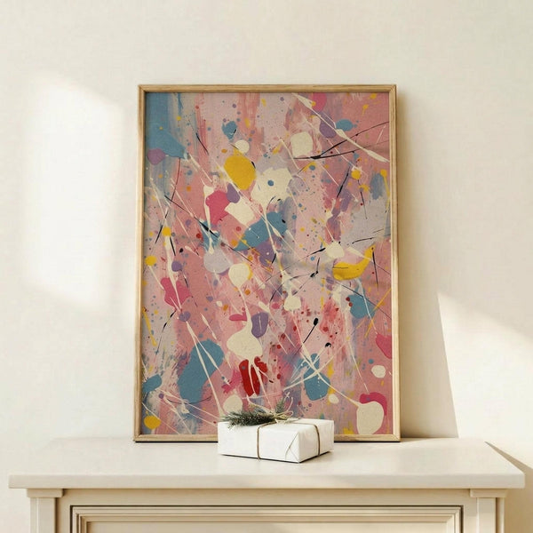 May include: Abstract expressionist style artwork with pink background featuring colorful paint splatters and drips in yellow, blue, red, and white with black lines throughout, displayed in thin wooden frame on light wood console table next to small wrapped white gift box with evergreen sprig, against cream wall