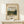 May include: Framed museum poster featuring Pissarro's landscape painting of rolling green hills, countryside village, and cloudy sky, displayed on light wood surface next to small wrapped gift with twine and evergreen sprig against cream wall