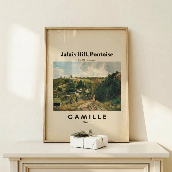 May include: Framed museum poster featuring Pissarro's landscape painting of rolling green hills, countryside village, and cloudy sky, displayed on light wood surface next to small wrapped gift with twine and evergreen sprig against cream wall