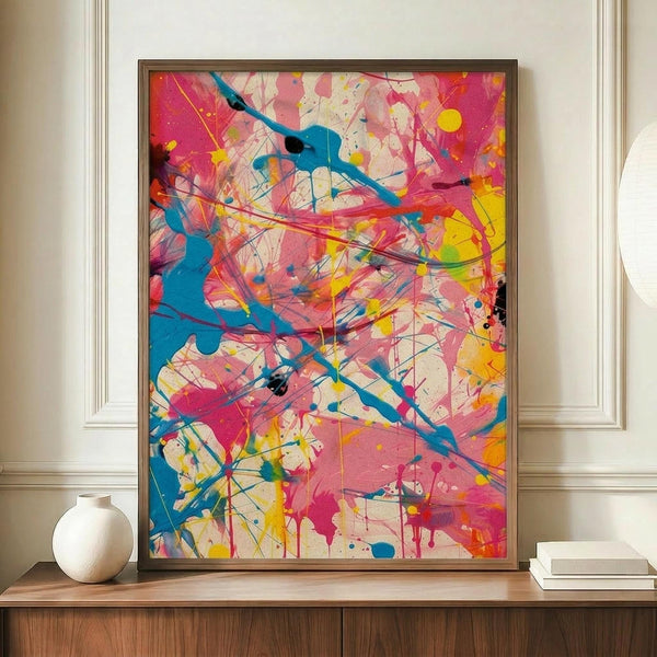 May include: Abstract expressionist artwork with vibrant paint splatters and drips in bright pink, turquoise blue, yellow, and orange against cream background. Framed print displayed on wooden sideboard next to white ceramic vase and stacked books in elegant room with cream walls and white molding.