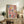 May include: Abstract expressionist artwork with vibrant paint splatters and drips in pink, blue, yellow, and red displayed in light wooden frame on rustic wood sideboard. Room scene shows fern plant, beige chair, ceramic vase, and stacked books creating warm, modern living space with neutral walls and natural lighting.