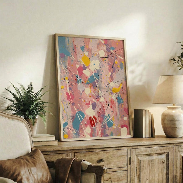 May include: Abstract expressionist artwork with vibrant paint splatters and drips in pink, blue, yellow, and red displayed in light wooden frame on rustic wood sideboard. Room scene shows fern plant, beige chair, ceramic vase, and stacked books creating warm, modern living space with neutral walls and natural lighting.