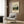 May include: Beige armchair with geometric pillow positioned next to gold floor lamp in modern living room. Framed vintage-style art poster featuring pastoral landscape with rolling green hills and countryside scene hangs on wood-paneled wall. Dark round side table with open book visible near window with white curtains.