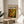 May include: Framed art poster featuring Jackson Pollock's abstract expressionist painting with vibrant green, yellow, and brown swirled patterns and white circular elements. The poster displays "CHAOS AND CREATIVITY 1912-1956 POLLOCK" text above the artwork. Shown in thin wooden frame, leaning against white wall on round wooden side table next to beige upholstered chair with plant leaves visible in soft natural lighting.