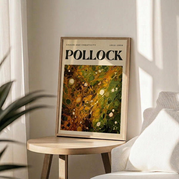 May include: Framed art poster featuring Jackson Pollock's abstract expressionist painting with vibrant green, yellow, and brown swirled patterns and white circular elements. The poster displays "CHAOS AND CREATIVITY 1912-1956 POLLOCK" text above the artwork. Shown in thin wooden frame, leaning against white wall on round wooden side table next to beige upholstered chair with plant leaves visible in soft natural lighting.