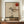 May include: Framed abstract artwork featuring a minimalist Japanese-inspired design with a solitary tree silhouette, red circle sun, and geometric black lines on neutral beige background. The poster is displayed on a wooden sideboard alongside a white ceramic vase and stacked books in a modern interior with cream walls and white molding.