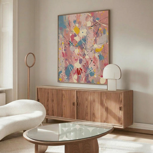 May include: Abstract splatter painting with vibrant pink, blue, yellow and purple paint drips and splatters on white background, displayed above wooden sideboard in modern living room with white curved chair, glass coffee table, and minimalist decor