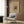 May include: Beige armchair with geometric patterned pillow in modern living room setting. Framed abstract art poster with colorful angular shapes and German text mounted on wood-paneled wall. Gold floor lamp, dark side table with open book, and neutral curtains complete the contemporary interior scene.