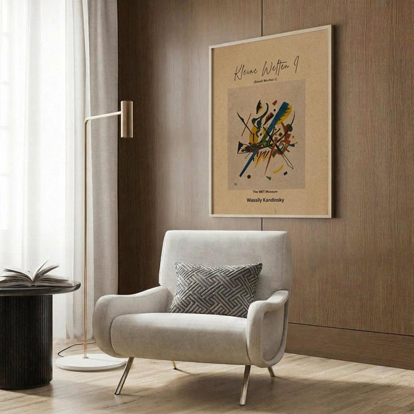 May include: Beige armchair with geometric patterned pillow in modern living room setting. Framed abstract art poster with colorful angular shapes and German text mounted on wood-paneled wall. Gold floor lamp, dark side table with open book, and neutral curtains complete the contemporary interior scene.
