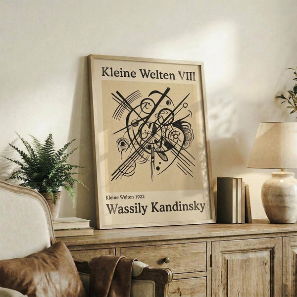 May include: Framed abstract art print featuring black geometric lines, circles, and organic shapes on beige background, displayed on rustic wooden sideboard alongside fern plant, ceramic vase, and books in neutral-toned living room with cream upholstered furniture and soft natural lighting.
