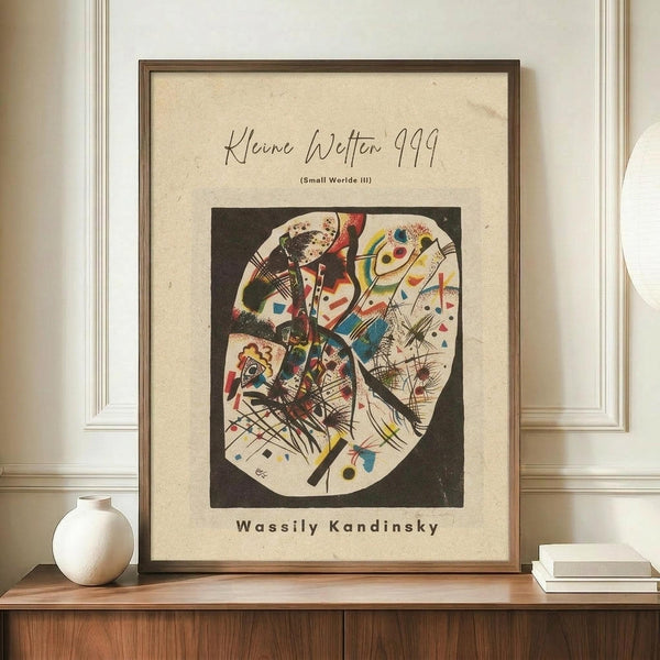 May include: Framed abstract art print featuring Wassily Kandinsky's colorful geometric composition with curved forms, lines, and shapes in red, blue, yellow, and black tones. The beige poster displays "Kleine Welten 999" and "Small Worlds III" text above the artwork. Shown in wooden frame on walnut credenza beside white ceramic vase and stacked books against cream wainscoted wall.