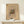 May include: Abstract art poster with colorful geometric shapes, lines and forms in yellow, blue, red and black on beige background, displayed in light wood frame on cream wooden console table next to small white wrapped gift with twine and evergreen sprig, against neutral wall with natural lighting.