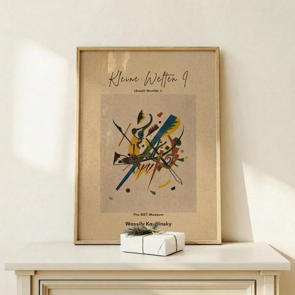 May include: Abstract art poster with colorful geometric shapes, lines and forms in yellow, blue, red and black on beige background, displayed in light wood frame on cream wooden console table next to small white wrapped gift with twine and evergreen sprig, against neutral wall with natural lighting.