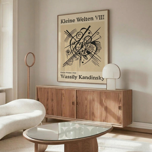 May include: A framed vintage-style poster featuring Wassily Kandinsky's abstract geometric artwork in black lines and curves on cream background, displayed above a mid-century modern wooden credenza with sliding doors. The room shows a white curved lounge chair, glass oval coffee table, modern brass floor lamp, and small white table lamp, creating a minimalist Scandinavian-inspired interior with neutral tones and natural lighting.