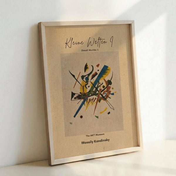May include: Framed abstract art print leaning against white wall on light wooden surface. Beige poster features colorful geometric shapes, lines and forms in yellow, blue, red, green and black. Artwork shows dynamic intersecting elements in modernist style. Natural wood frame surrounds the print. Soft natural lighting creates gentle shadows on wall.