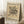 May include: Framed abstract art print leaning against white paneled wall on textured cream rug. Features geometric black line drawing with intersecting curves, circles, and radiating lines on beige background. German text at top and bottom with artist name. Light wood frame, positioned next to small wooden block with white coffee cup and saucer. Soft natural lighting creates gentle shadows.