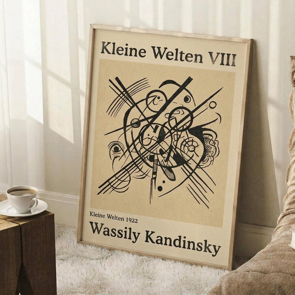 May include: Framed abstract art print leaning against white paneled wall on textured cream rug. Features geometric black line drawing with intersecting curves, circles, and radiating lines on beige background. German text at top and bottom with artist name. Light wood frame, positioned next to small wooden block with white coffee cup and saucer. Soft natural lighting creates gentle shadows.